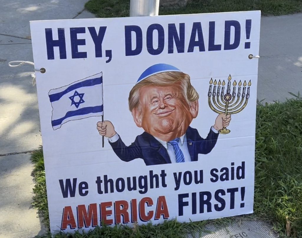 a sign showing former President Donald Trump waving an Israeli flag in one hand anda  Hanukah menorah in the other while wearing a kippah--a traditional Jewish headcovering worn during prayer. Text reads "Hey, Donald: We thought you said AMERICA FIRST" Sign is posted outside a synagogue in Ann Arbor, MI