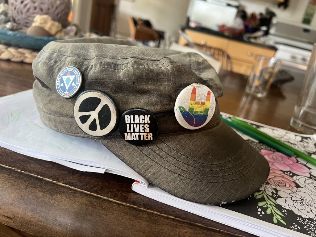A fairly beat-down green brimmed baseball cap with four buttons on it. The buttons show: 1) a "love" hand in rainbow colors, 2) the text "BLACK LIVES MATTER", 3) a peace symbol, and 4) a stylized Jewish star and the text "Secret Jewish Space Lasers Corps: Mazel Tough"