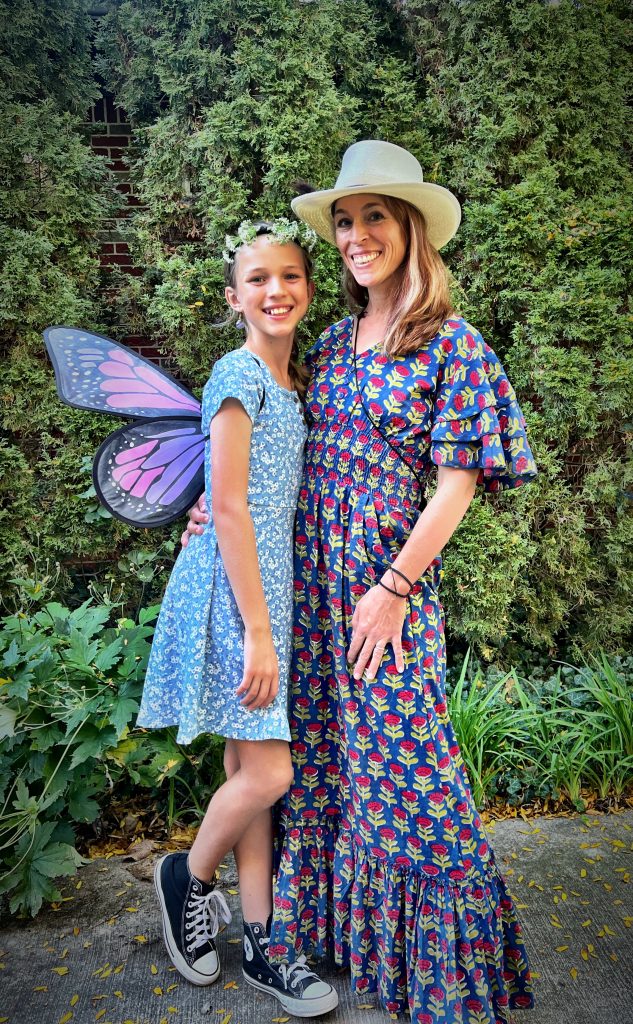 A woman and a girl standing in front of greenery, both wearing floral-patterned dresses. The girl has butterfly wings and a flower crown, while the woman is wearing a wide-brimmed hat.
