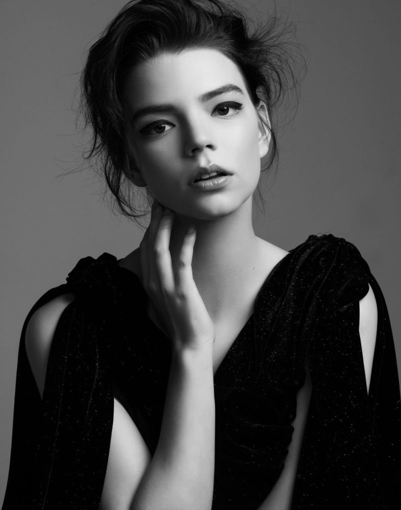 A black and white portrait of Anya Taylor-Joy with her hand on her neck. She is wearing a dark dress.